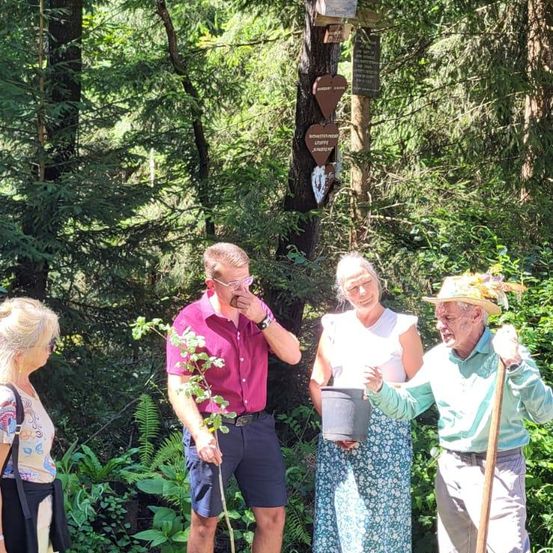 Vier Personen stehen in einem bewaldeten Bereich, umgeben von üppiger Vegetation. Ein Mann in einem lila Hemd hält eine Pflanze, während ein anderer Mann mit Hut einen Eimer in der Hand hat. Eine Frau in einem Blumenkleid lächelt. Hinter ihnen ist ein hölzerner Baumstamm mit herzförmigen Dekorationen zu sehen.