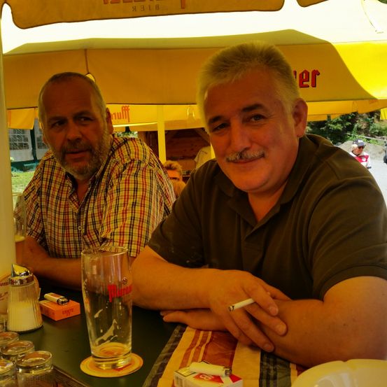 Zwei Männer sitzen unter einem gelben Regenschirm. Ein Mann hat eine Zigarette und ein Glas Bier. Ein anderer Mann neben ihm mit einem karierten Hemd. Dahinter ist ein verschwommener Hintergrund von einem Zelt und Bäumen.