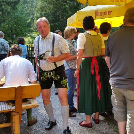 Eine Gruppe von Menschen in traditioneller bayerischer Kleidung versammelt sich im Freien unter einem gelben Regenschirm. Ein Mann hält ein Bierglas und eine Frau steht neben ihm in einem grünen Kleid.
