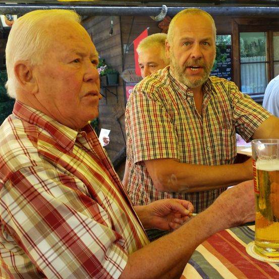 Zwei ältere Männer sitzen an einem Tisch, einer hält ein Glas Bier. Der Mann rechts gestikuliert mit der Hand. Hinter ihnen steht ein weiterer Mann in der Nähe einer Wand mit einer Fahne.