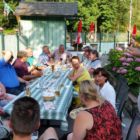 Eine Gruppe von Menschen sitzt um einen Tisch herum und genießt Getränke und Essen im Freien. Eine Frau lächelt im Vordergrund, während eine Fahne über einem kleinen Gebäude weht.