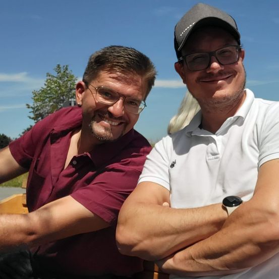 Zwei Männer sitzen nebeneinander, einer in einem Weinroten Hemd und der andere in einem weißen Poloshirt, beide lächeln und posieren für die Kamera.