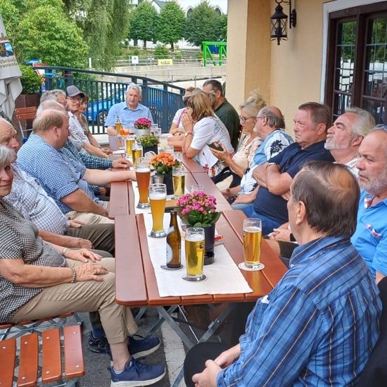 Eine Gruppe von Menschen sitzt draußen um einen Tisch herum. Sie alle lächeln und scheinen eine gute Zeit zu haben. Der Tisch ist mit Biergläsern, Flaschen und Blumen gefüllt. Im Hintergrund befinden sich Bäume und ein Zaun.