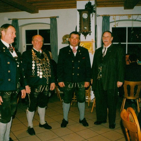 Vier Männer stehen in einem Raum, gekleidet in traditioneller Kleidung. Zwei tragen Medaillen, und einer hält eine Zigarette. Eine Uhr hängt an der Wand, und zwei Personen sitzen an einem Tisch im Hintergrund.