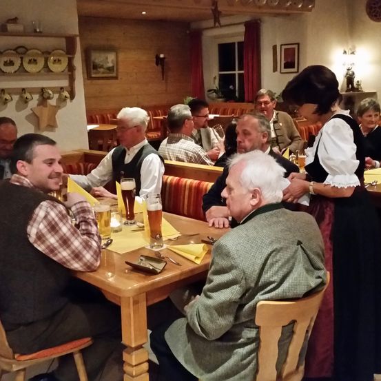 Mehrere Männer sitzen an einem Tisch in einem Restaurant und trinken Bier. Sie sind von anderen Gästen umgeben. Eine Frau in traditioneller Kleidung steht hinter einem Mann in einem grünen Jackett.