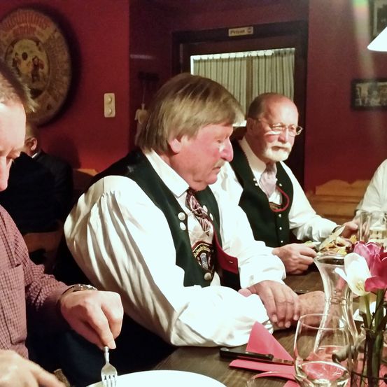 Drei Männer in traditioneller Kleidung sitzen um einen Esstisch, einer von ihnen hält eine Gabel. Der Tisch hat eine Vase mit rosa Blumen und Weingläser. Der Raum hat rote Wände und eine Holztür.