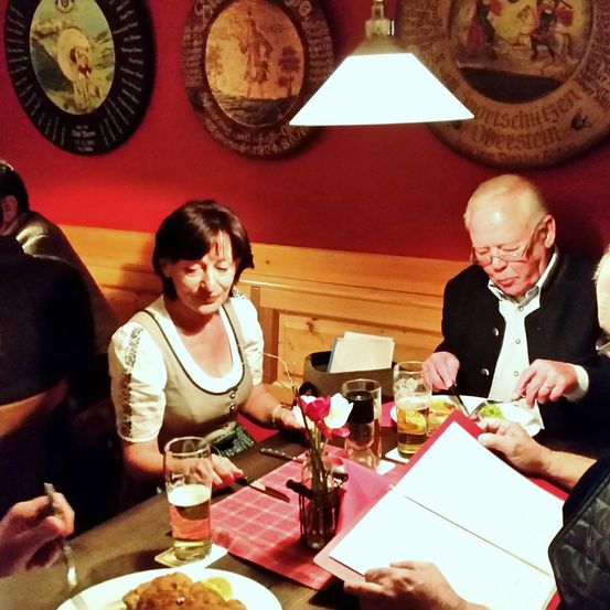 Eine Gruppe älterer Menschen isst in einem Restaurant und trägt traditionelle Kleidung. Der Tisch hat eine Speisekarte, Getränke und eine Blumenvase. Die Wände sind mit dekorativen Tellern verziert.