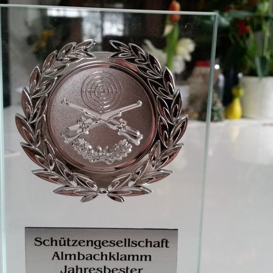 Eine Glastrophäe mit einem silbernen Emblem, das zwei gekreuzte Gewehre und ein Ziel zeigt, mit der Aufschrift 'Schützengesellschaft Almbachklamm Jahresbester'.