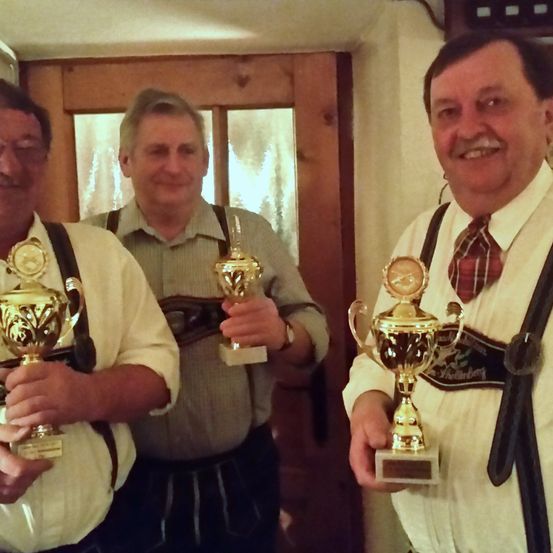 Drei Männer in traditioneller deutscher Kleidung halten Pokale. Sie stehen vor einer Holztür.