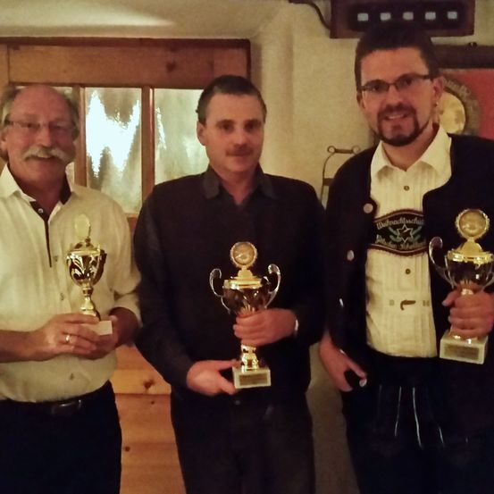 Drei Männer in einem Raum, die Pokale halten und für ein Foto lächeln. Der Mann auf der linken Seite trägt ein weißes Hemd und hat einen Schnurrbart. Der Mann in der Mitte trägt ein schwarzes Hemd und hat eine Brille. Der Mann auf der rechten Seite trägt einen schwarzen Anzug und hat einen Bart.