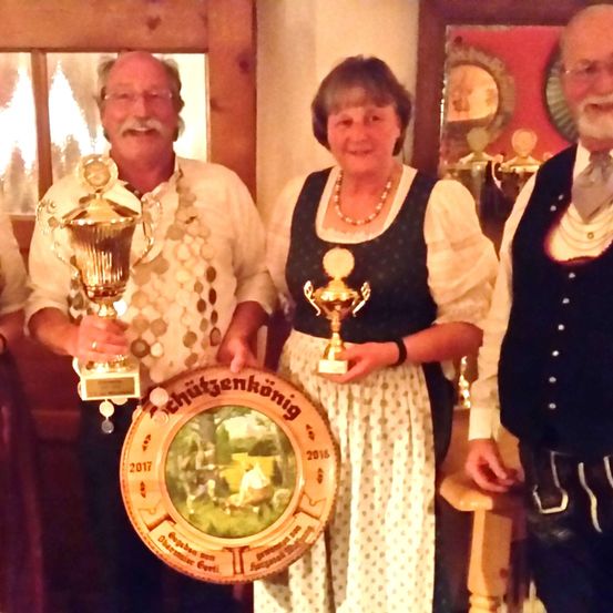 Vier Menschen in traditioneller Kleidung stehen eng beieinander und halten Trophäen. Die Frau in der Mitte hält eine runde Auszeichnung und die anderen haben einzelne Trophäen. Sie stehen vor einer Holztür.