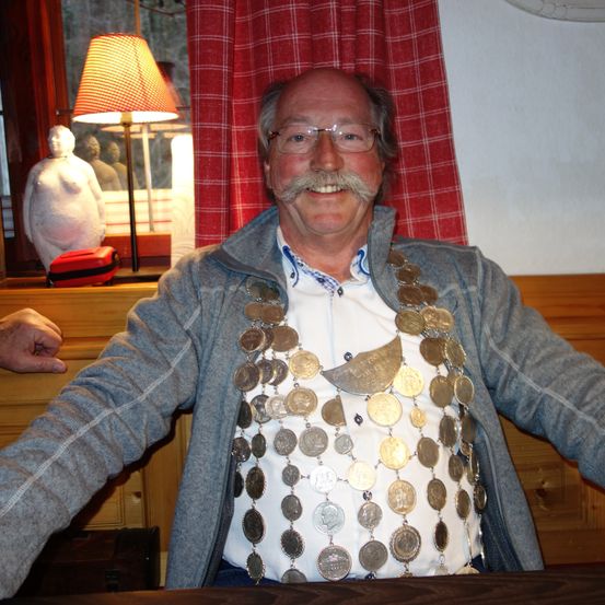 Ein älterer Mann mit einem Schnurrbart und Brille lächelt und trägt eine graue Jacke und eine Halskette aus Goldmünzen. Hinter ihm hängt ein rot-weiß karierter Vorhang, und eine Lampe steht auf einem Fensterbrett.