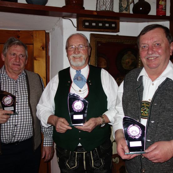 Drei Männer in traditioneller Kleidung stehen nebeneinander, lächeln und halten Auszeichnungen. Hinter ihnen ist eine Holztür und Regale mit dekorativen Gegenständen zu sehen.