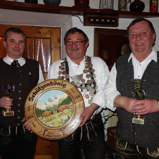 Drei Männer stehen nebeneinander, lächeln und halten Trophäen. Der Mann in der Mitte trägt eine dekorative Münzkette und hält eine große runde Holzplatte mit einem Bild von Rehen.