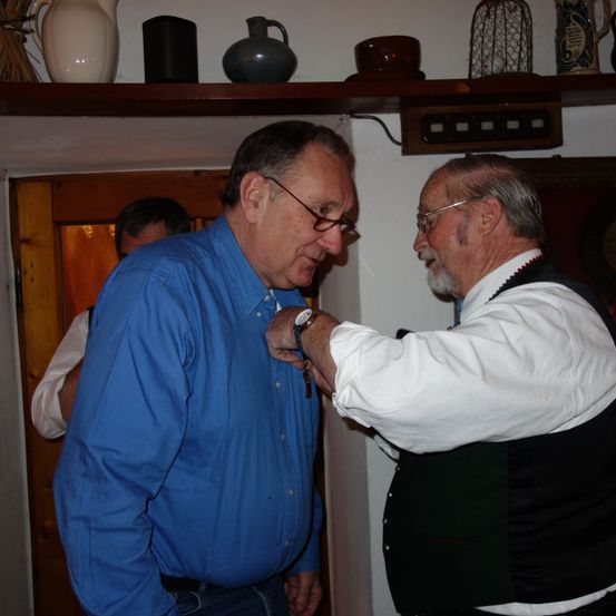 Zwei ältere Männer unterhalten sich in einem Raum mit einer Holztür, einer trägt ein blaues Hemd, der andere ein weißes Hemd mit einem schwarzen West. Dahinter steht ein Regal mit verschiedenen Gegenständen.