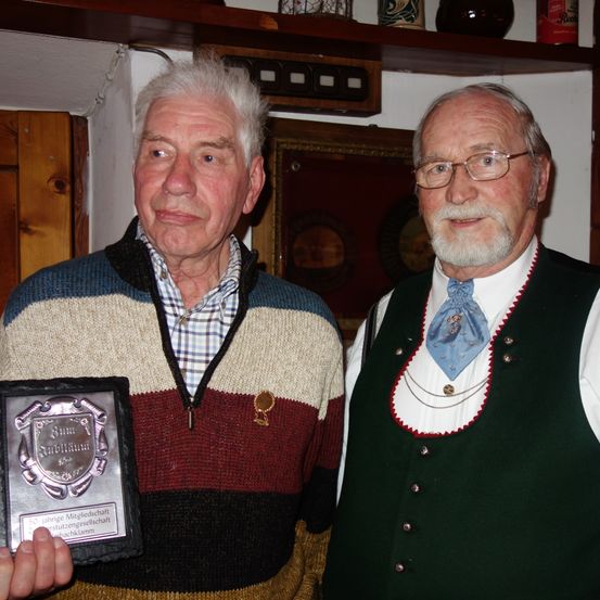Zwei ältere Männer stehen nebeneinander. Einer hält eine Plakette mit der Aufschrift 'Baum Schuller'. Beide tragen traditionelle Kleidung, wobei der rechte eine Brille und eine blaue Weste trägt. Hinter ihnen steht ein Holzregal mit verschiedenen Gegenständen.