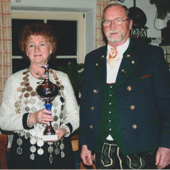 Ein Mann und eine Frau stehen zusammen in einem Raum. Die Frau hält eine Trophäe. Der Mann trägt eine Brille und traditionelle Kleidung.