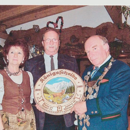 Drei Personen in traditioneller Kleidung, möglicherweise bei einer Feier, halten eine Plakette mit einem Hirsch und Bergen. Der Mann auf der rechten Seite trägt eine Medaille und eine Kette.