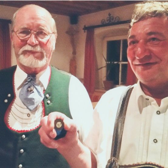 Zwei Männer, beide lächelnd, posieren zusammen. Der Mann auf der linken Seite trägt eine grüne Weste und eine Fliege. Der Mann auf der rechten Seite hält einen Zigarre. Hinter ihnen ist ein Fenster mit einem roten Vorhang.