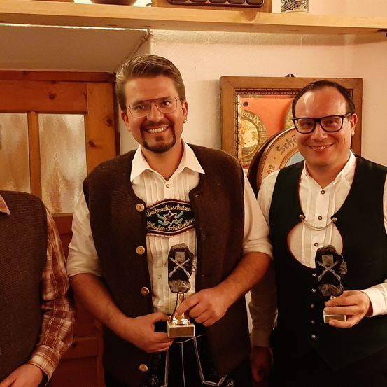 Drei Männer in traditioneller Kleidung halten Trophäen und stehen in einem Raum mit Holzwänden und Regalen. Sie lächeln für die Kamera.