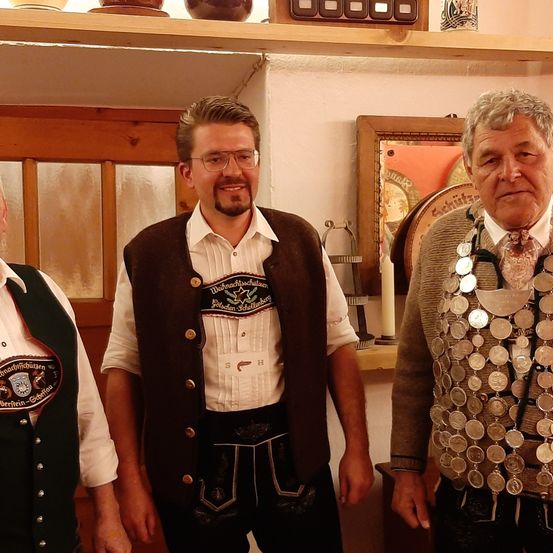 Drei Männer stehen eng beieinander in einem Raum. Der Mann in der Mitte trägt eine braune Weste mit einem weißen Hemd und einer Brille. Der Mann auf der rechten Seite trägt einen grauen Pullover mit einer Münzkette.