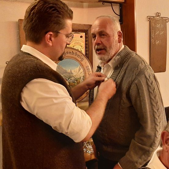 Zwei Männer stehen eng beieinander, einer befestigt eine Medaille am anderen. Der Mann, der befestigt, trägt eine Brille und eine Weste, während der Medaillenträger einen grauen Pullover hat. Hinter ihnen ist ein gerahmtes Bild mit Bergen und einem Kalender.