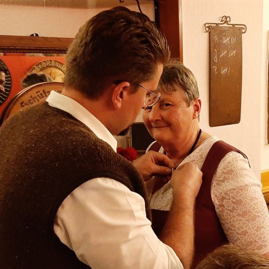 Ein Mann mit Brille befestigt eine Brosche an einer lächelnden Frau in traditioneller Kleidung. Sie befinden sich in einem Raum mit einem Holzrahmen und einer weißen Wand mit Text.