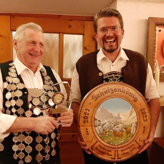 Zwei Männer stehen in einem Raum, beide lächeln und halten Auszeichnungen. Ein Mann hält eine Plakette mit einer Bergszene und Text, während der andere einen Pokal hält. Beide sind in traditioneller Kleidung gekleidet.