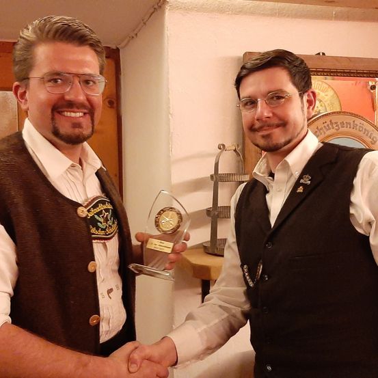 Zwei Männer in traditioneller Kleidung lächeln und schütteln sich die Hände vor einer Holzwand. Einer von ihnen hält einen Pokal. Dahinter steht ein Holzschrank mit einer dekorativen Platte.