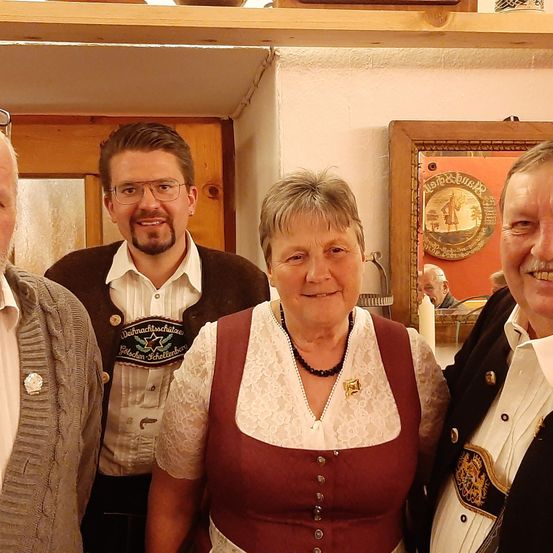 Drei Personen, zwei Männer und eine Frau, stehen lächelnd zusammen. Der Mann auf der linken Seite trägt einen grauen Pullover, während der mittlere Mann eine braune Jacke und ein weißes Hemd trägt. Die Frau trägt ein kastanienbraunes Kleid mit Spitze. Hinter ihnen befindet sich eine Holztür, ein Regal und ein Spiegel.