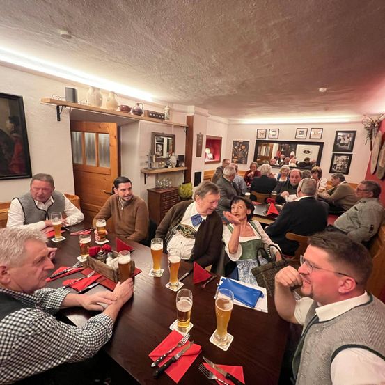 Eine Gruppe von Menschen sitzt um einen Tisch in einem Restaurant mit Biergläsern, Tellern und Besteck. An der Wand hängen mehrere Bilderrahmen.