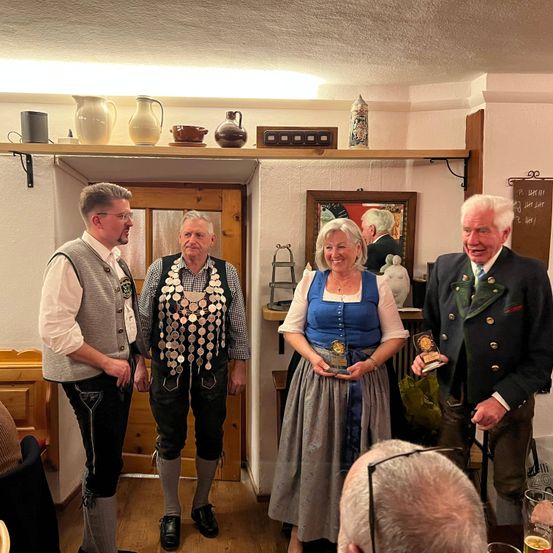 Vier Personen in traditioneller deutscher Kleidung stehen lächelnd nebeneinander und halten Auszeichnungen. Sie befinden sich in einem Raum mit einer Holztür und einem Regal mit Dekorationsartikeln. Ein Spiegel und eine Tafel mit Schriftzug hängen an der Wand.