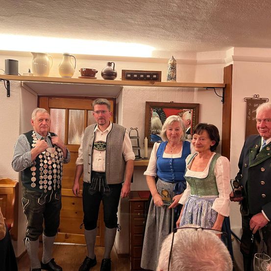 Fünf Personen, zwei Männer und drei Frauen, in traditioneller Kleidung, stehen zusammen in einem Raum. Sie scheinen für ein Foto zu posieren. Hinter ihnen ist eine Holztür, ein gerahmtes Bild und ein Regal mit verschiedenen Gegenständen zu sehen.