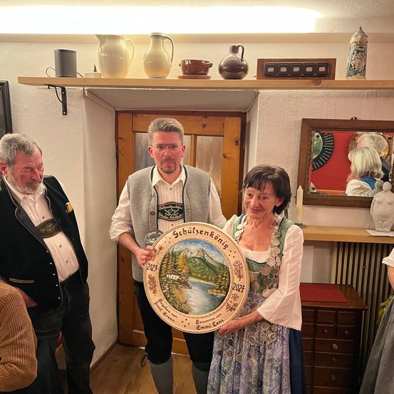 Zwei Männer und eine Frau in traditioneller Kleidung stehen in einem Raum. Der Mann rechts hält einen kreisförmigen Schild. Hinter ihnen befindet sich ein Holzregal mit verschiedenen Gegenständen.