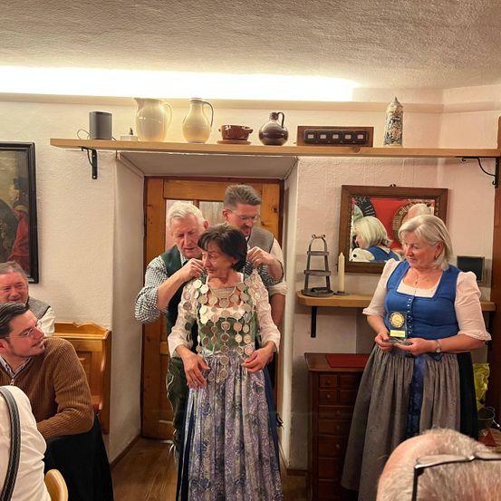 Eine Gruppe von Menschen in einem Raum, eine ältere Frau wird von einem Mann unterstützt, während eine weitere Frau in traditioneller Kleidung steht. Der Raum ist mit verschiedenen Gegenständen und Kunstwerken dekoriert.