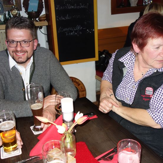 Ein Mann und eine Frau sitzen in einem Restaurant an einem Tisch und posieren für ein Foto. Auf dem Tisch stehen Gläser mit Bier, eine rote Serviette und Blumen. Hinter ihnen steht eine Tafel mit Schrift.