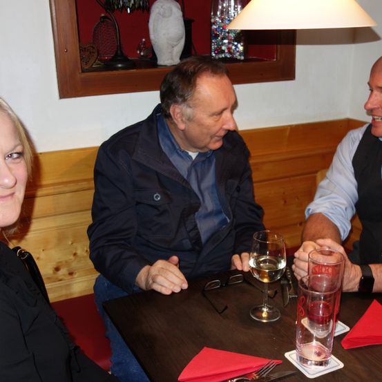 Drei Personen sitzen an einem Restauranttisch, zwei Männer und eine Frau. Der Mann auf der linken Seite trägt eine Jacke und der Mann auf der rechten Seite trägt eine Weste. Weinläser und Servietten sind auf dem Tisch.