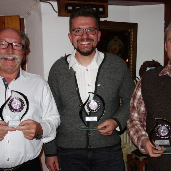 Drei Männer in einem Raum halten Pokale. Der Mann auf der linken Seite hat einen Schnurrbart, der Mann in der Mitte lächelt und der Mann auf der rechten Seite trägt ein kariertes Hemd.