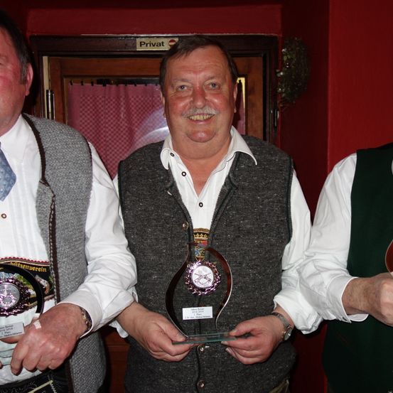 Drei Männer stehen vor einer roten Wand. Der Mann in der Mitte hält eine Trophäe mit einem Uhrdesign. Der Mann auf der linken Seite hält eine Plakette. Der Mann auf der rechten Seite lächelt.