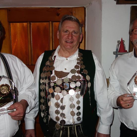 Drei Männer stehen nebeneinander. Der Mann in der Mitte trägt eine Münzkette und eine Weste. Der Mann auf der linken Seite trägt eine Hosenträger und hält eine Trophäe. Der Mann auf der rechten Seite trägt eine Brille und hält eine Trophäe.