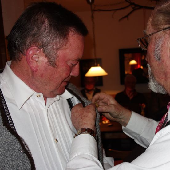 Ein älterer Mann in einem weißen Hemd und einem grauen Pullover wird von einem anderen älteren Mann mit einer Medaille ausgezeichnet. Die Medaillenverleihung findet in einem schwach beleuchteten Raum mit hängenden Lampen und einem Spiegel statt.