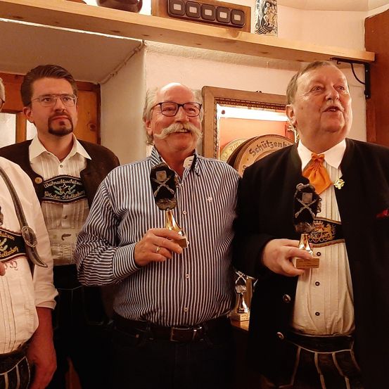 Vier Männer in traditioneller deutscher Kleidung halten Trophäen in der Hand und stehen in einem Raum mit einem Holzregal über ihnen. Der Mann in der Mitte hat einen Schnurrbart und eine Brille.