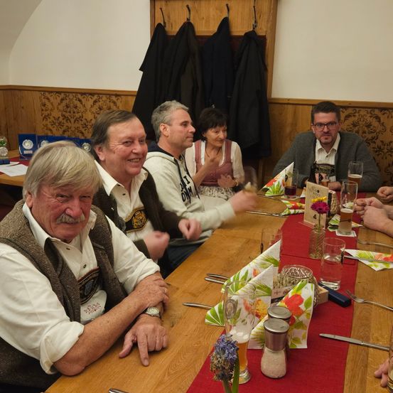 Eine Gruppe älterer Erwachsener sitzt an einem langen Tisch in einem Restaurant, lächelt und genießt ihre Getränke und Speisen. Ein roter Tischläufer bedeckt den Tisch, und verschiedene Gerichte und Getränke sind vor ihnen aufgestellt.