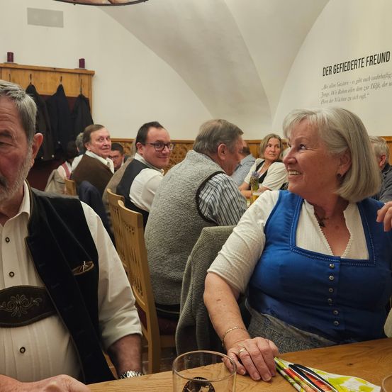 Eine Gruppe von Menschen, darunter ein älteres Paar, sitzt in einem Essbereich. Sie tragen traditionelle Kleidung. Die Frau lächelt und schaut in die Kamera. Vor ihnen steht ein Tisch mit einem Glas und Besteck. Dahinter befindet sich eine Wand mit einem Zitat in deutscher Sprache.