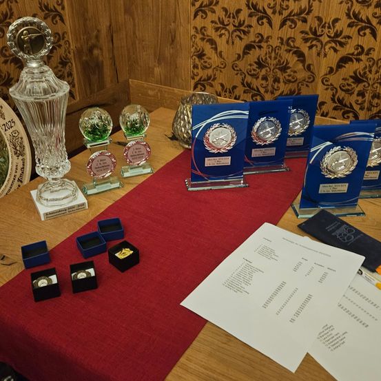 Ein hölzerner Tisch mit einem roten Tischläufer zeigt Trophäen und Auszeichnungen, darunter eine große Kristalltrophäe, kleinere Glastrophäen und blaue Plaketten mit silbernen Medaillen. Verschiedene kleine Schachteln mit Medaillen und ein Stück Papier mit Text sind ebenfalls vorhanden.