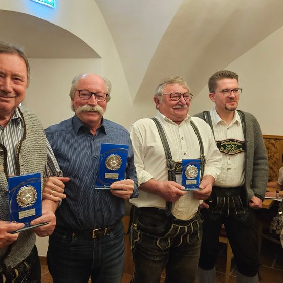 Vier Männer stehen eng beieinander, lächelnd, und halten Auszeichnungen in den Händen. Die Männer tragen Freizeitkleidung und Brillen. Ein Mann trägt eine Hose mit Hosenträgern. Der Mann rechts trägt eine Uhr. Es gibt einen Holztisch mit einigen Gegenständen darauf.