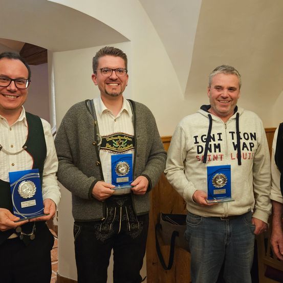 Vier Männer stehen zusammen, lächeln und halten Auszeichnungen, zwei in traditioneller deutscher Kleidung, einer in einer Kapuzenjacke mit 'IGNITION DAM MAP SAINT LOUIS' und einer in einem weißen Hemd.