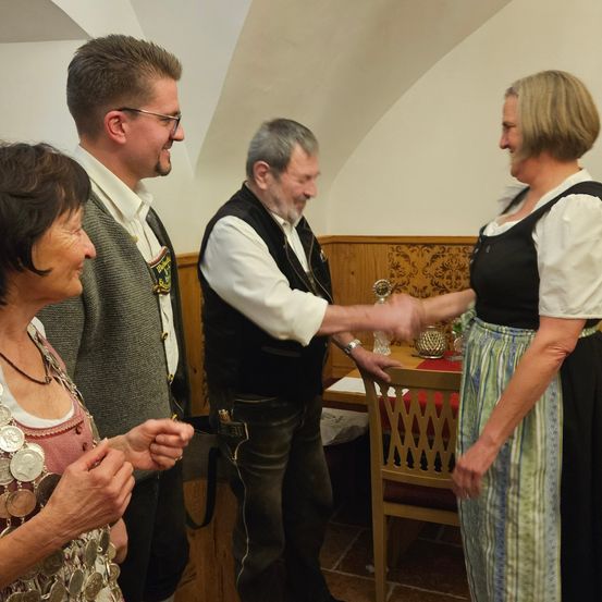 Vier Menschen stehen in einem Raum, zwei Frauen und zwei Männer. Eine Frau schüttelt einem Mann die Hand, während die andere Frau eine Münzkette hält. Der Raum hat Holzwände und einen Holztisch.