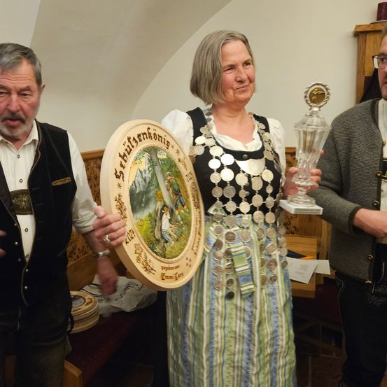 Eine Frau hält eine große Holzplakette und einen Glaspokal, flankiert von zwei Männern in traditioneller Kleidung, möglicherweise bei einer Zeremonie oder Preisverleihung.