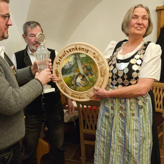 Eine Frau in einer traditionellen Tracht mit vielen Münzen um die Taille hält einen dekorativen Teller, während zwei Männer neben ihr stehen, einer mit einem Glaspokal. Sie scheinen sich bei einer Veranstaltung zu befinden, möglicherweise bei einer Preisverleihung.